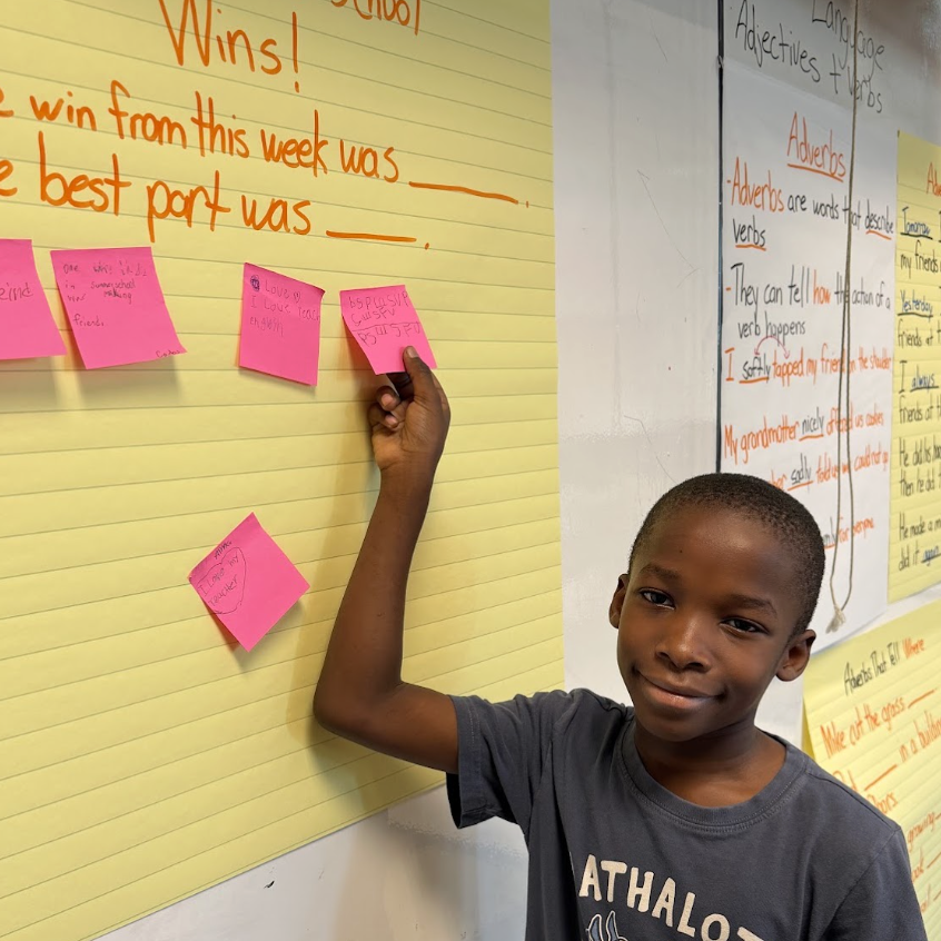 A young boy smiles while holding a pink sticky note on a yellow lined chart in a classroom.