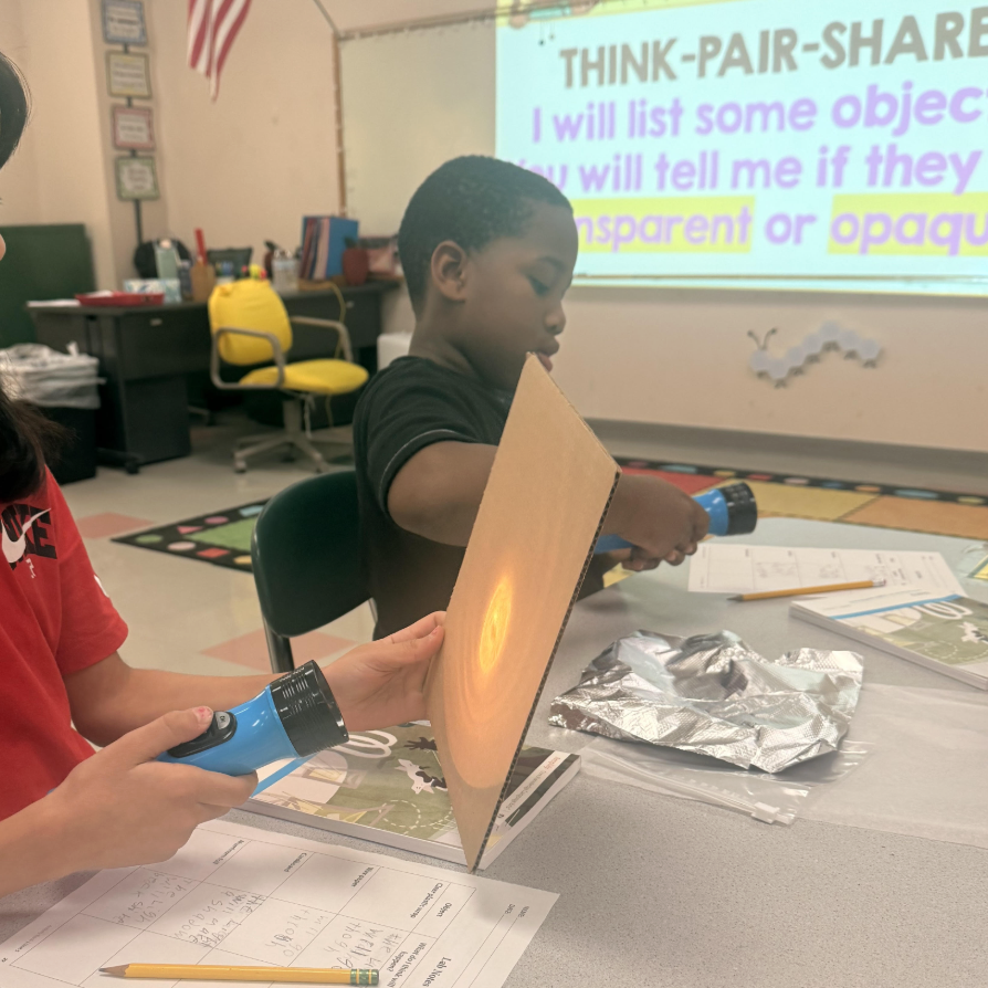 Students conduct a science experiment in a classroom, using flashlights and cardboard to test transparency.