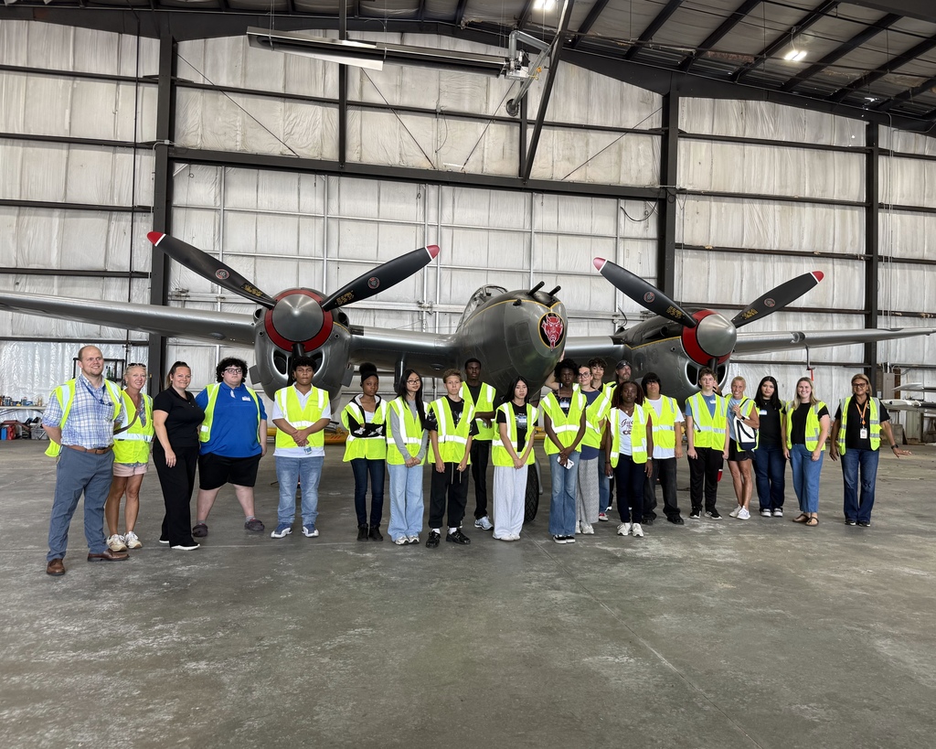 A group of students in visibility vests stand in front of an airplane at the Worcester City Airport.