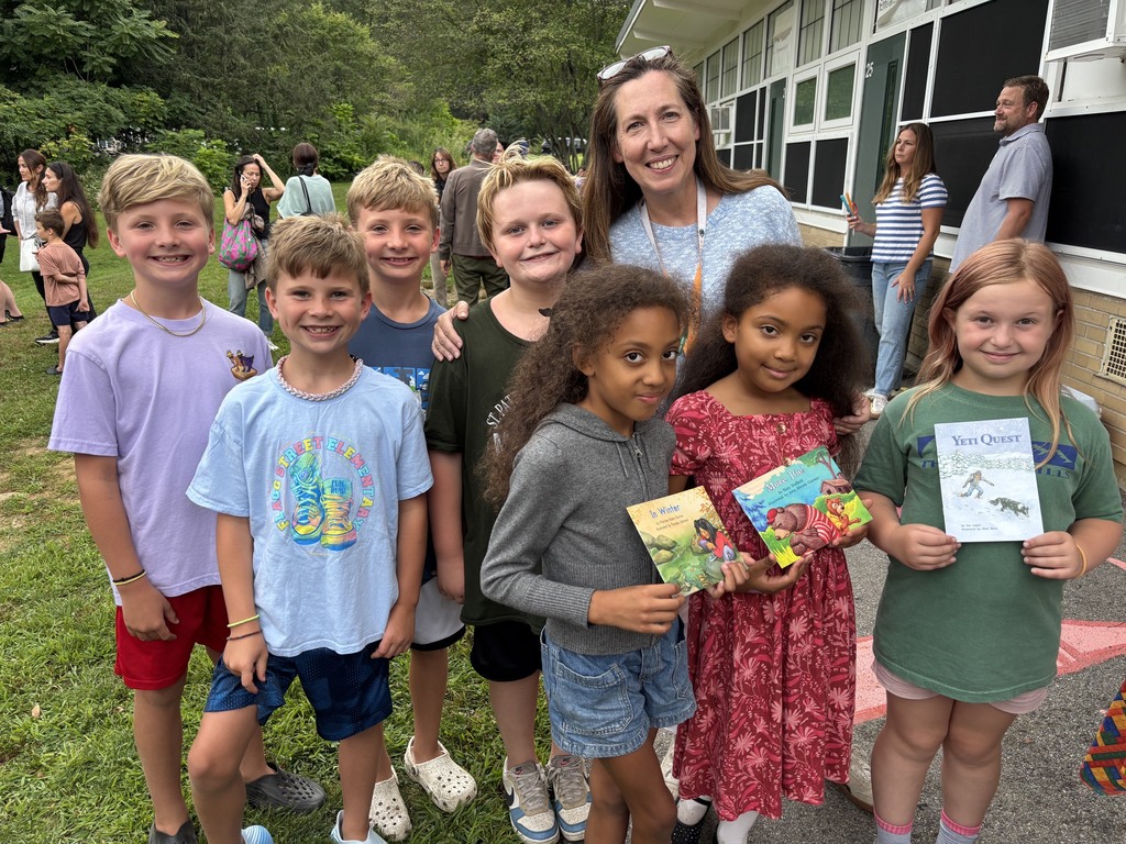 A group of smiling children and an adult stand together outdoors, some holding books.