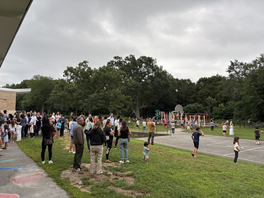A group of people, including adults and children, gather outdoors near a school building and playground.