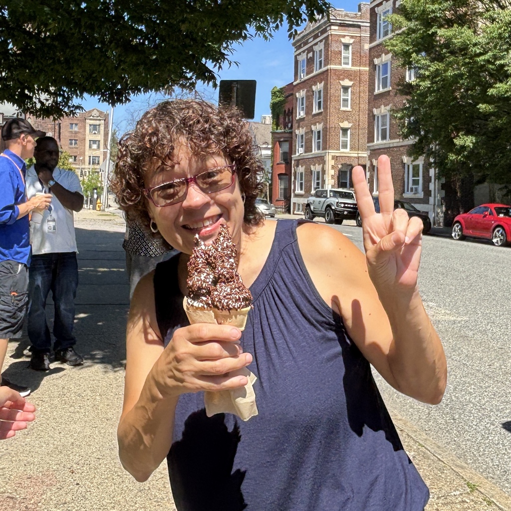 A woman smiles while holding a chocolate ice cream cone with sprinkles and making a peace sign.