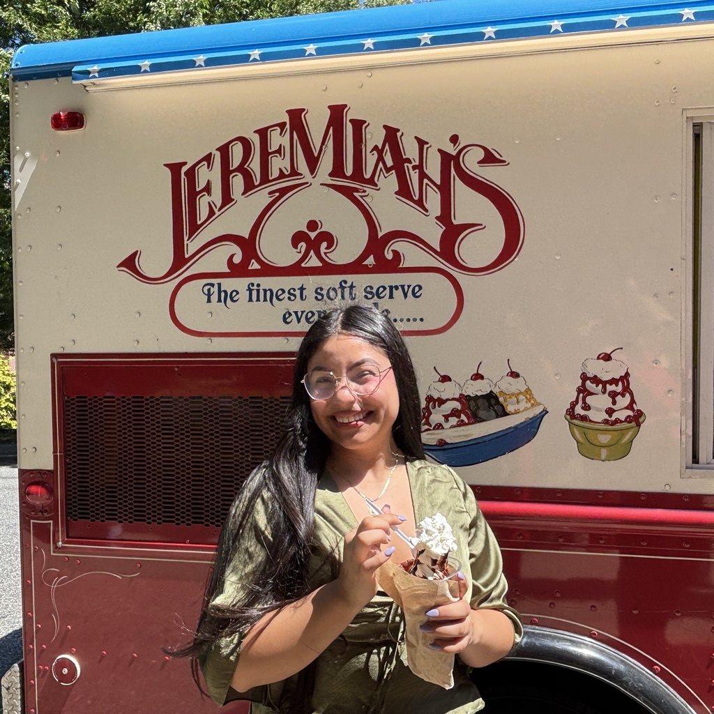A smiling young person holds a cup of soft-serve ice cream in front of a Jeremiah's ice cream truck.