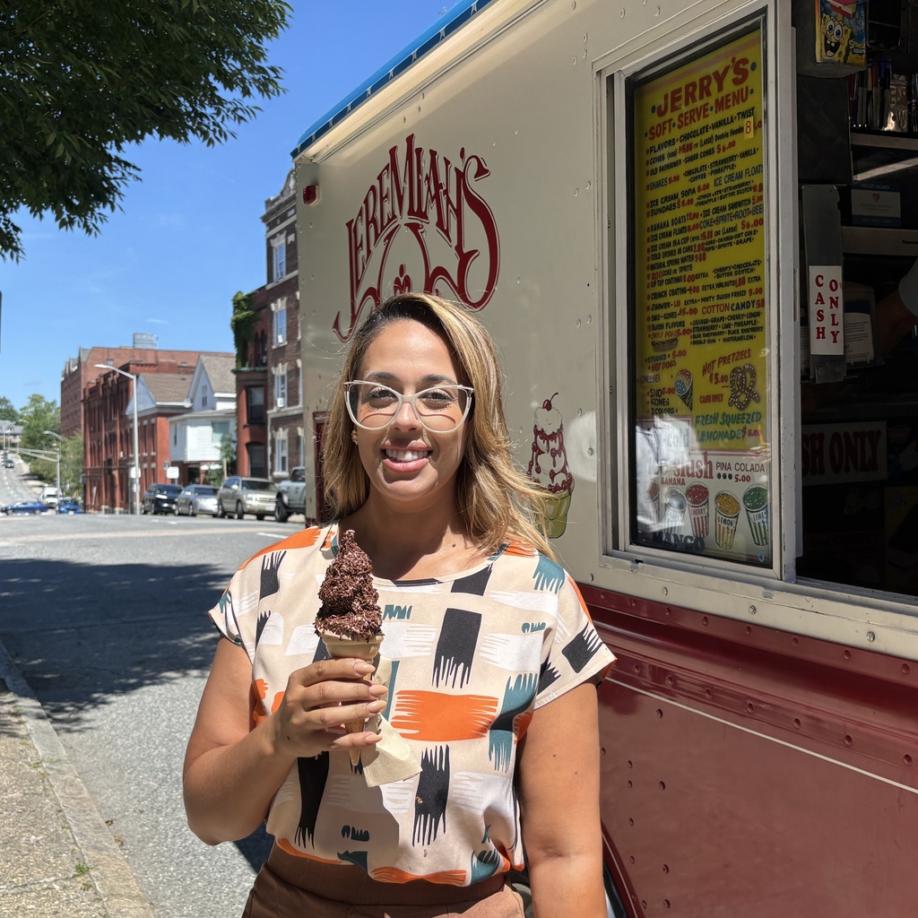 A smiling woman stands on a sidewalk holding a chocolate soft-serve ice cream cone in front of an ice cream truck