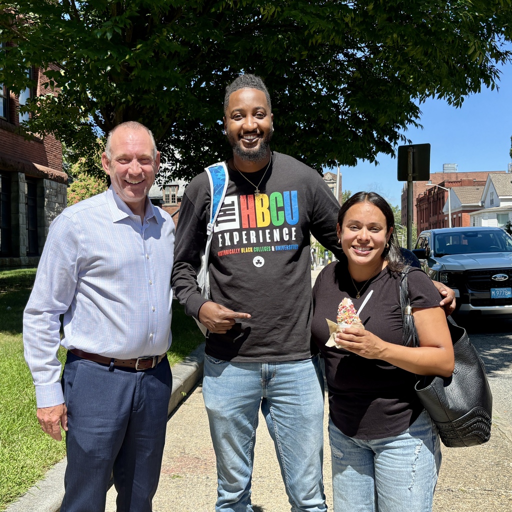 Three people stand together outdoors, smiling in front of a brick building and trees.