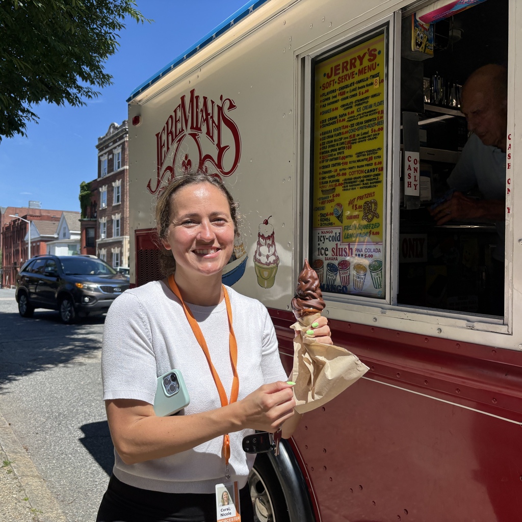 A smiling woman holds a chocolate-dipped ice cream cone in front of an ice cream truck with a menu visible.
