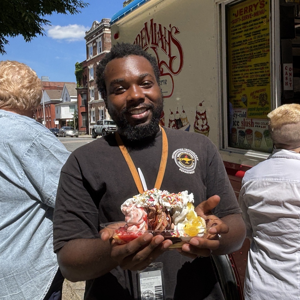 A smiling man holds a large ice cream sundae with whipped cream and sprinkles in front of an ice cream truck.