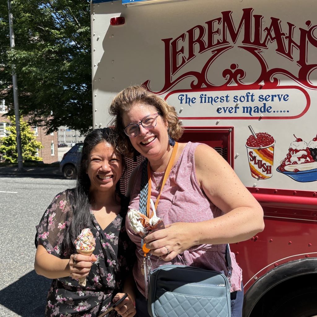 Two smiling women stand in front of an ice cream truck, each holding a frozen treat.