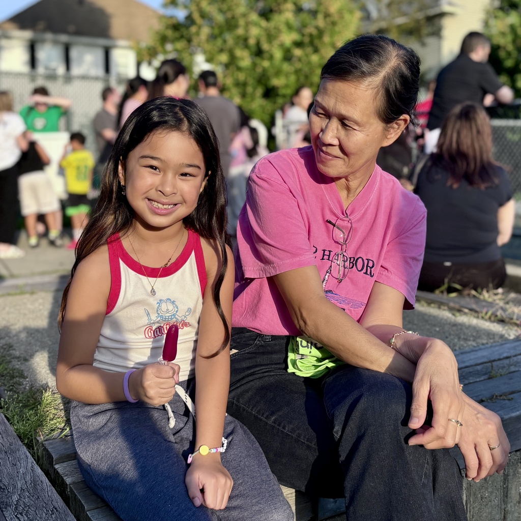 A young girl with dark hair smiles brightly while holding a red popsicle, sitting next to an adult woman.