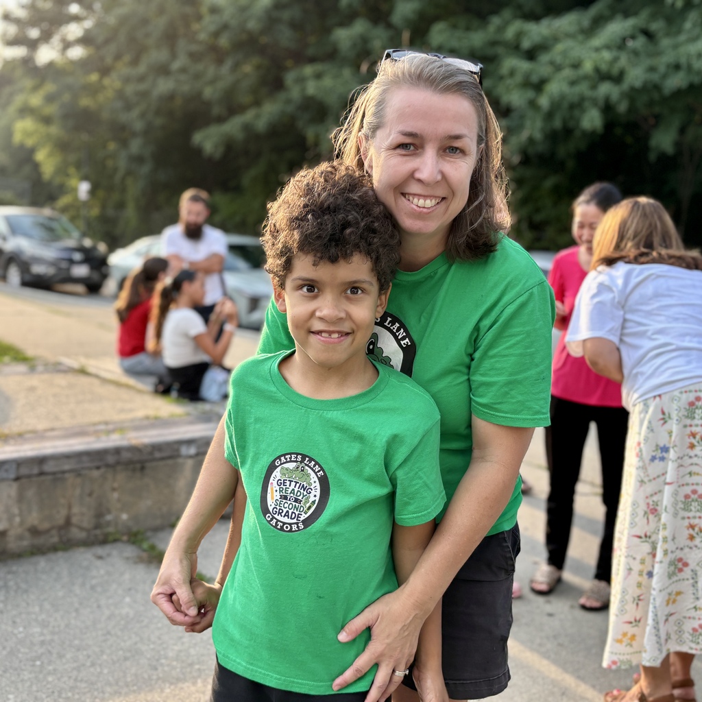 A woman and a young boy in matching green t-shirts smile at the camera, with other people in the background.