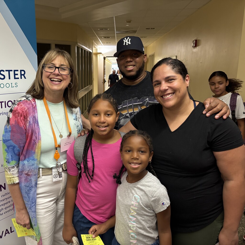 A diverse group of people, including a family and a school principal, smile for a photo in a hallway.