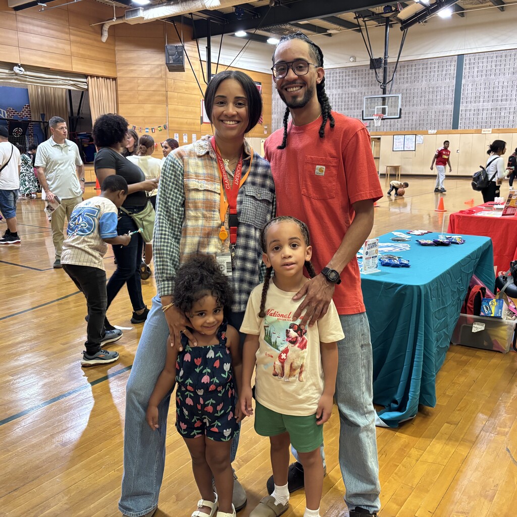 A smiling family of four poses for a photo in a gymnasium with other people in the background.