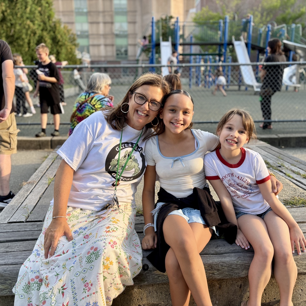 A woman and two girls smile while sitting on a wooden bench at a playground.
