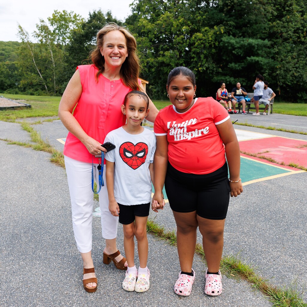 A woman smiles with two young girls, one wearing a Spider-Man shirt and the other a red shirt with text.