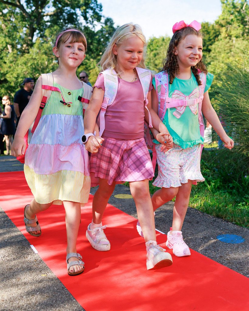 Three young students hold hands while walking on a red carpet. 