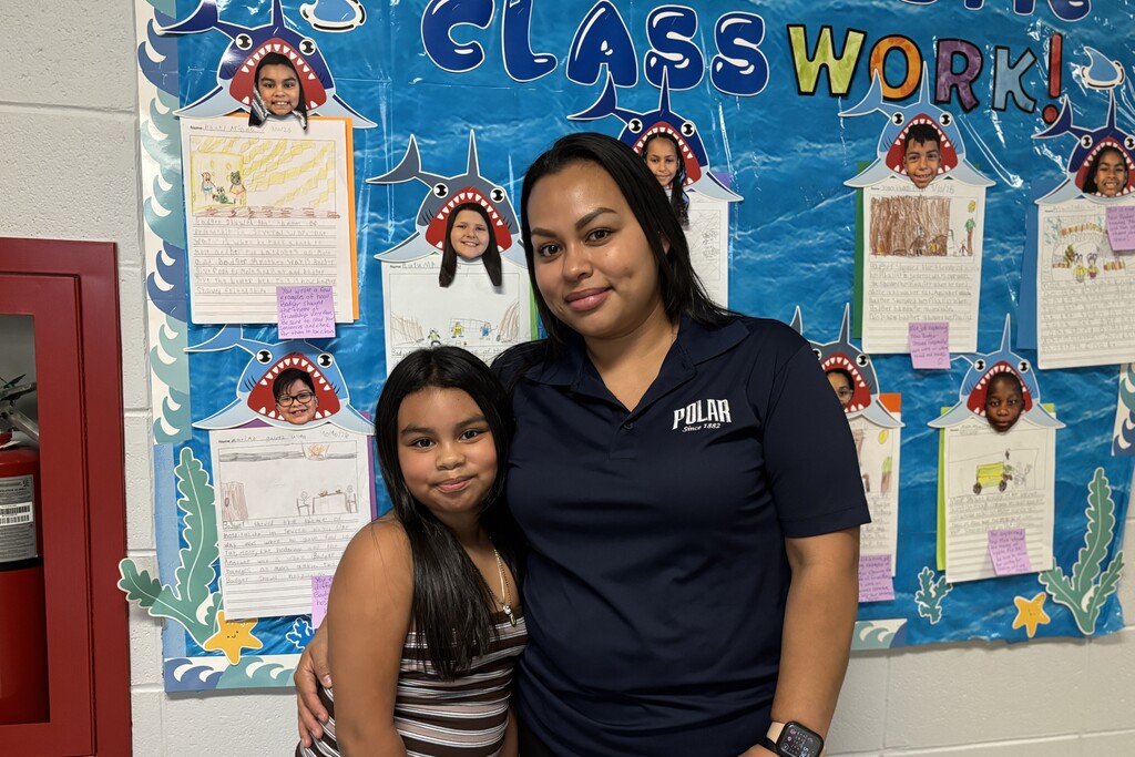 A teacher and a student stand together in front of a blue classroom bulletin board decorated with shark cutouts and student artwork.