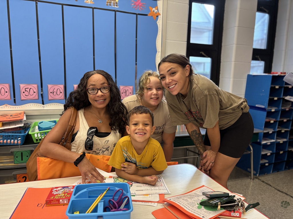 A group of four smiles around a desk in a colorful classroom, with worksheets and school supplies on the table.