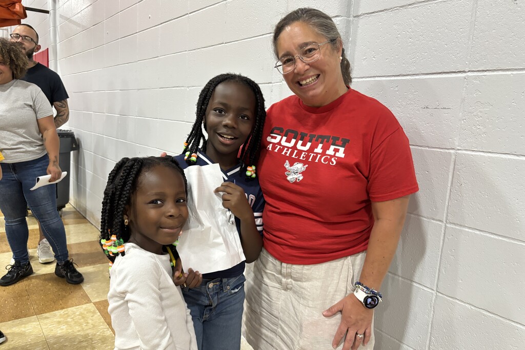 A smiling woman wearing a red "South Athletics" shirt poses with two young girls against a white brick wall.
