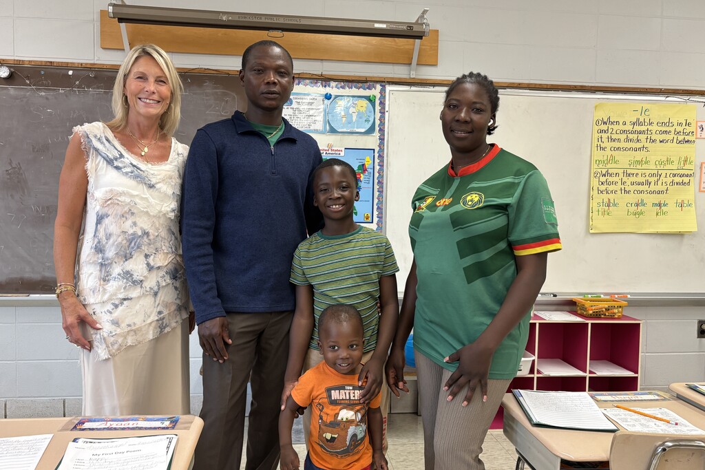 A family of four stands smiling in a classroom next to a teacher.