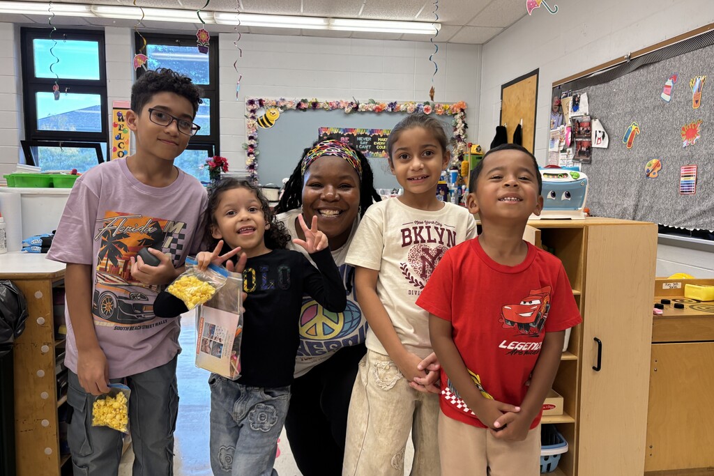 A teacher and four smiling students pose together in a brightly lit classroom.