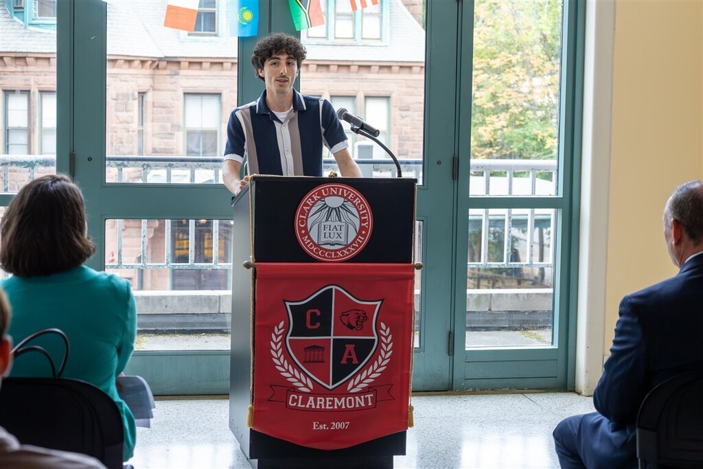 A young person with curly dark hair speaks at a lectern featuring a Claremont banner and a Clark University seal.