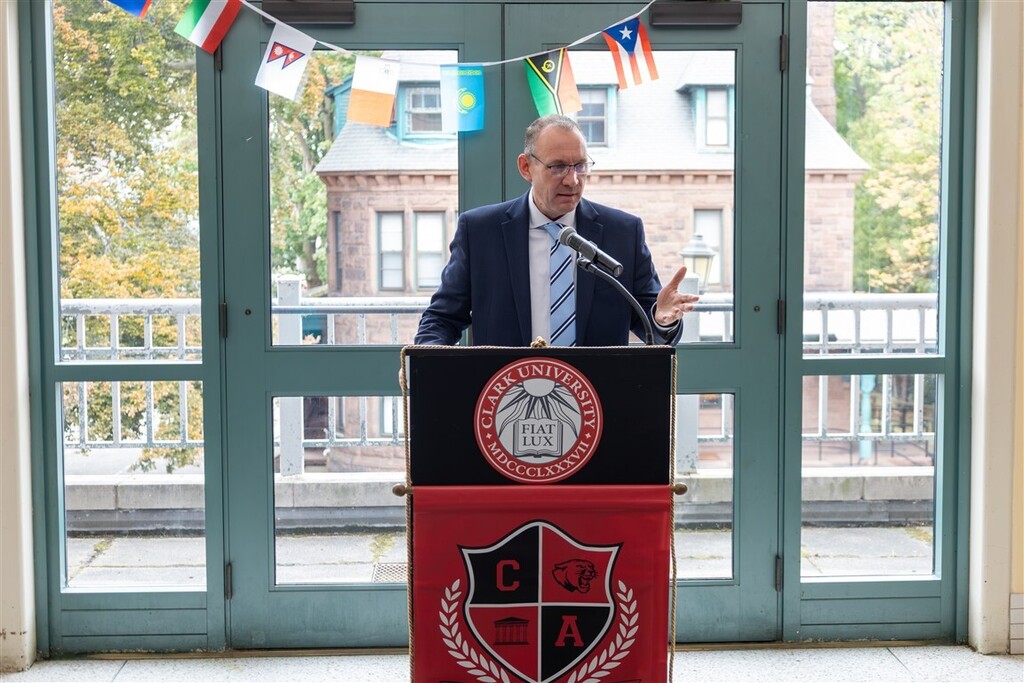 A man in a suit speaks at a Clark University podium decorated with international flags.