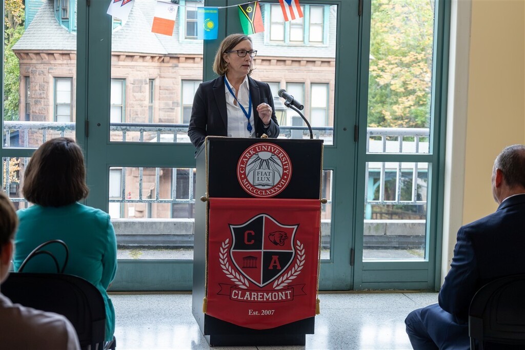 A woman wearing glasses and a blazer speaks at a podium with a 'Clark University Claremont' seal, in front of an audience.
