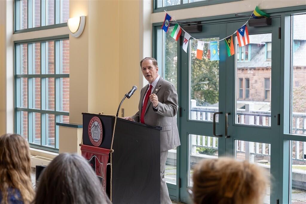 A speaker in a suit stands behind a podium with the Clark University seal, addressing an audience in a sunlit room decorated with international flags.