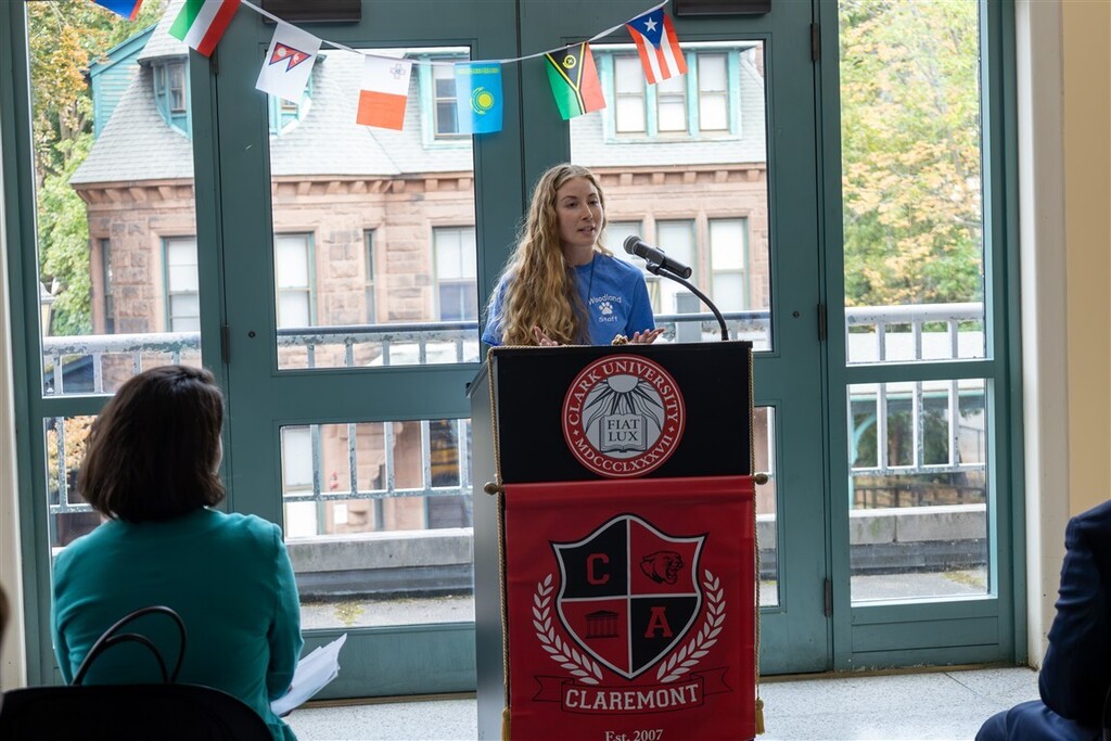 A young woman with long blonde hair speaks at a lectern with the Clark University and Claremont logos, addressing an audience.