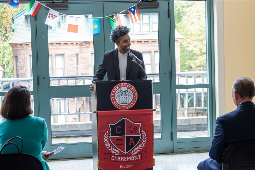 A woman wearing glasses and a blazer speaks at a podium with a 'CLARK UNIVERSITY' seal and a banner that reads 'CLAREMONT Est. 2007'.