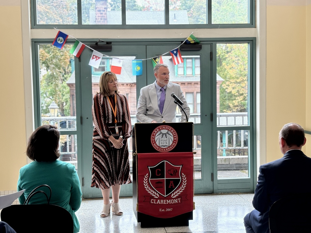 A man speaks at a podium with the Clark University Claremont logo, while a woman stands beside him and an audience listens indoors.