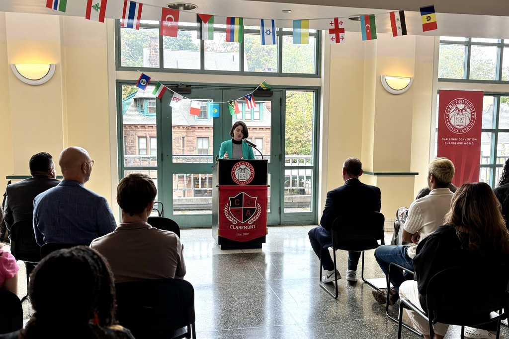 A young person with curly dark hair speaks at a podium with the Clark University and Claremont seals.