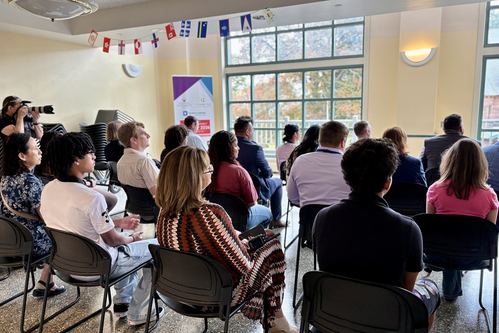 A diverse group of people sits in chairs, attentively listening to a presentation in a sunlit room with large windows.