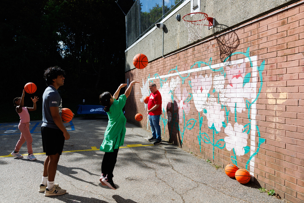 A young student shoots a basketball at the hoop while volunteers observe nearby.