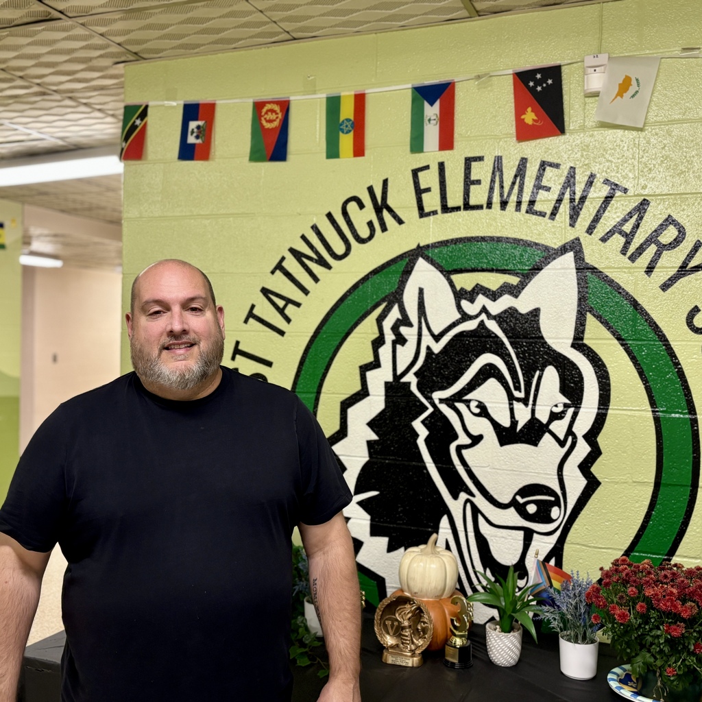 A man in a black t-shirt stands before a school mural featuring a husky and the text "WEST TATNUCK ELEMENTARY".