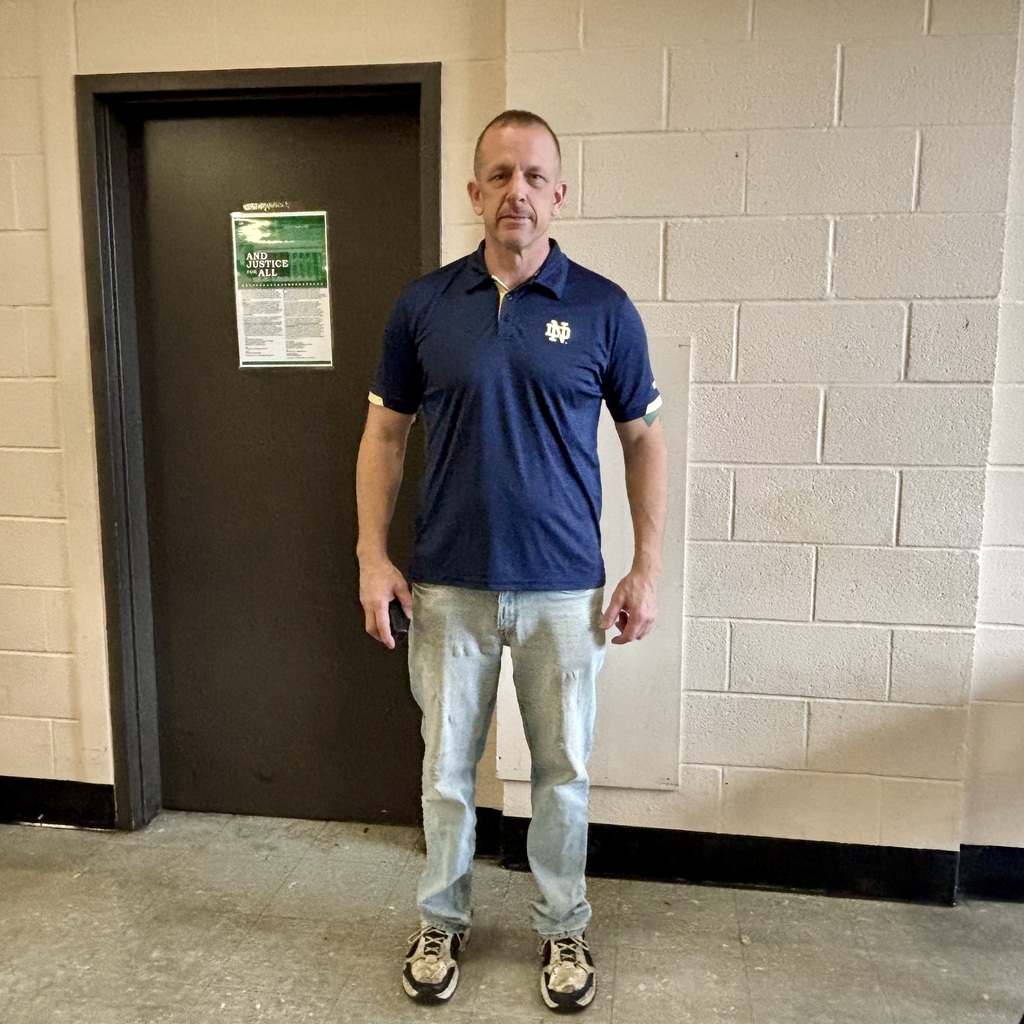 A man in a blue polo shirt and jeans stands in a hallway in front of a closed door with a green sign.