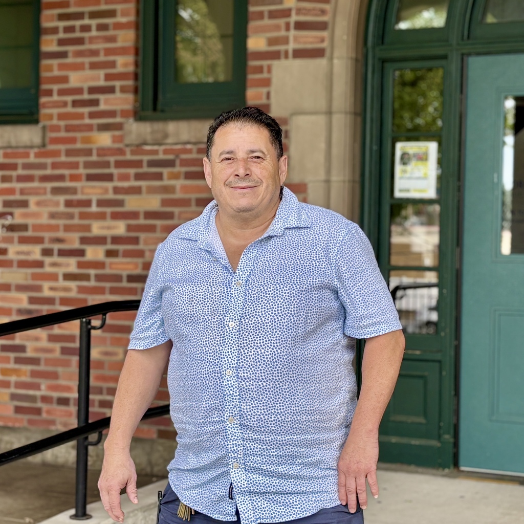 A smiling man in a blue patterned shirt stands outside a brick building with green doors.