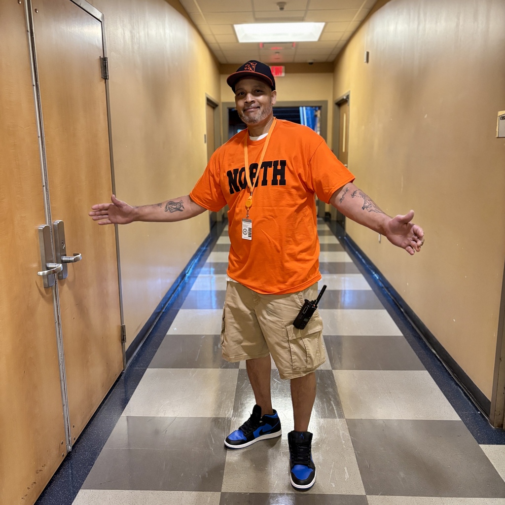 A person wearing a bright orange 'NORTH' t-shirt, cap, and beige shorts stands in a well-lit hallway with arms outstretched.