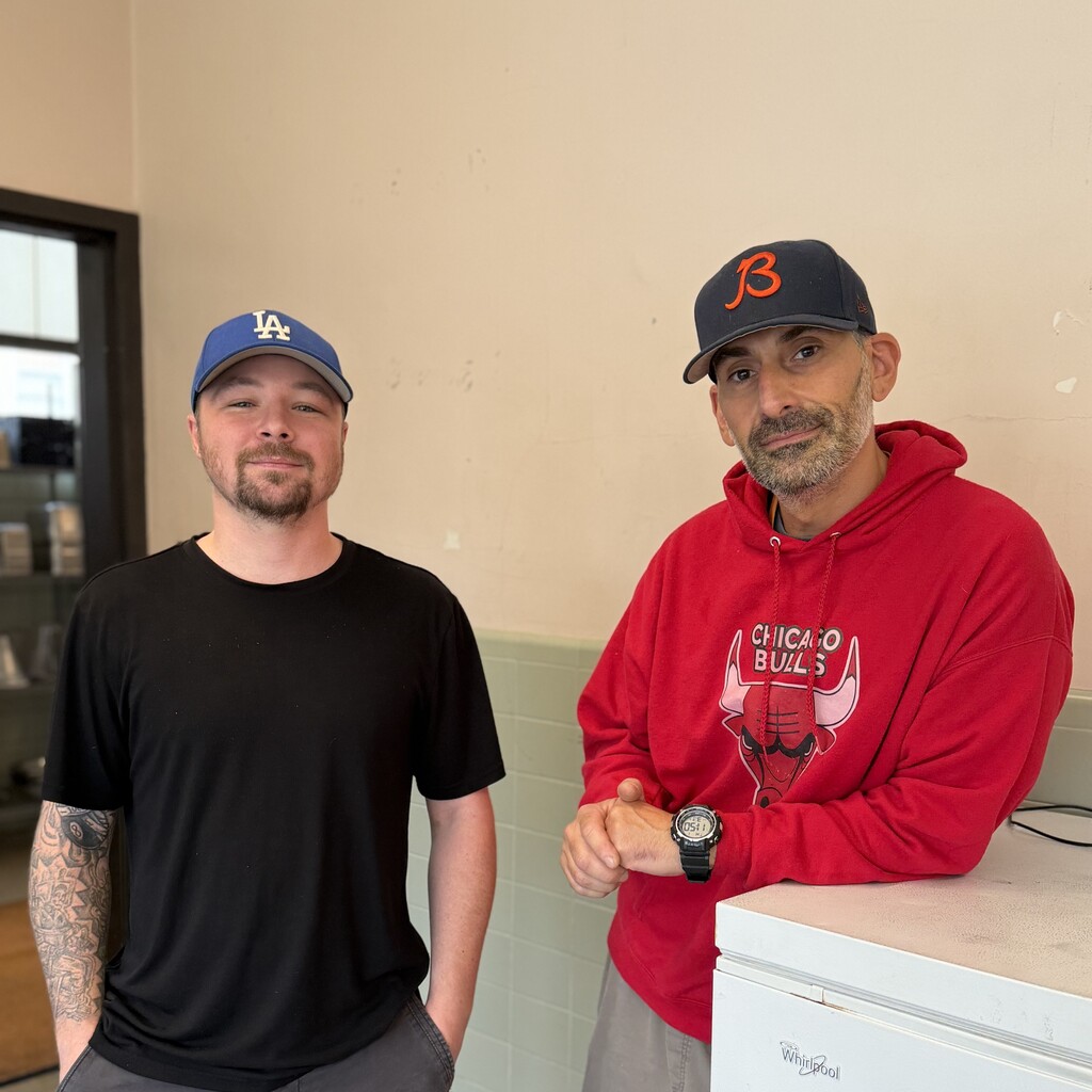 Two men stand side-by-side indoors, wearing baseball caps and casual clothing.