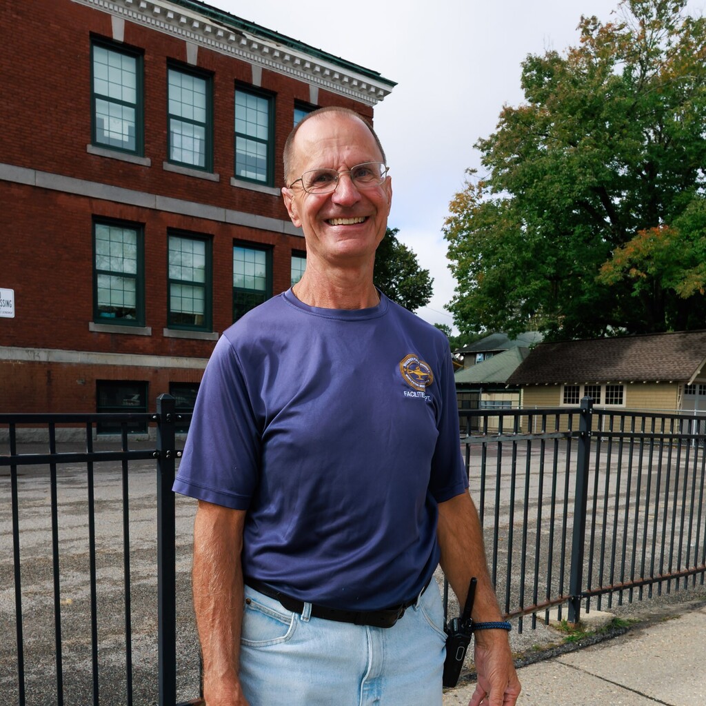 A smiling man wearing glasses and a blue shirt stands outdoors in front of a brick building.