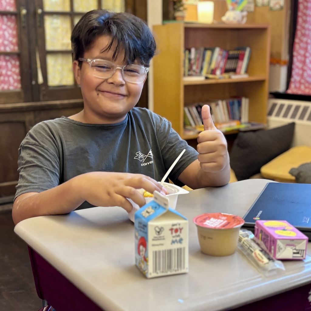 A smiling girl sits at a school desk, holding a cup of applesauce with a spoon.