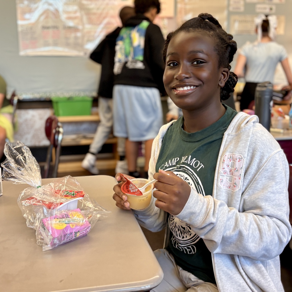 A smiling girl sits at a school desk, holding a cup of applesauce with a spoon.