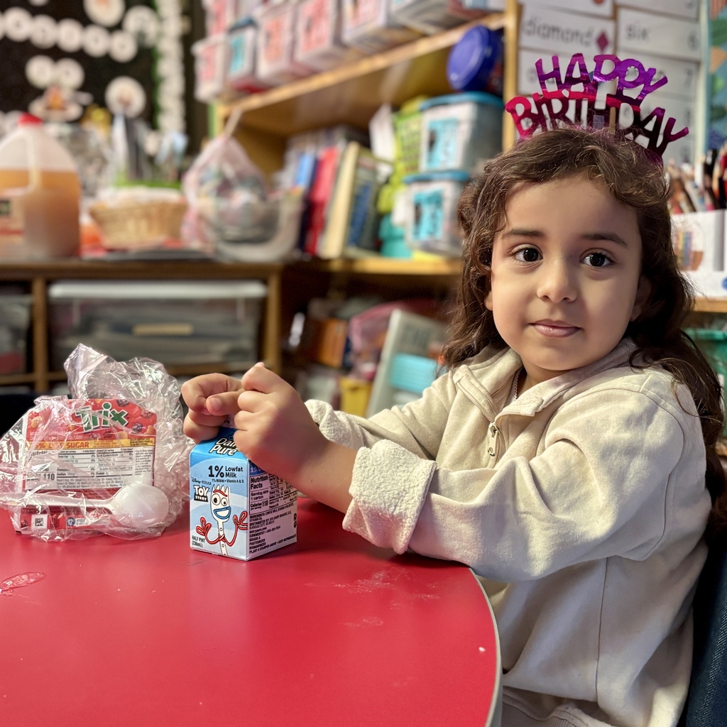 A child wearing a sparkly birthday tiara sits at a red table with a milk carton and a snack.