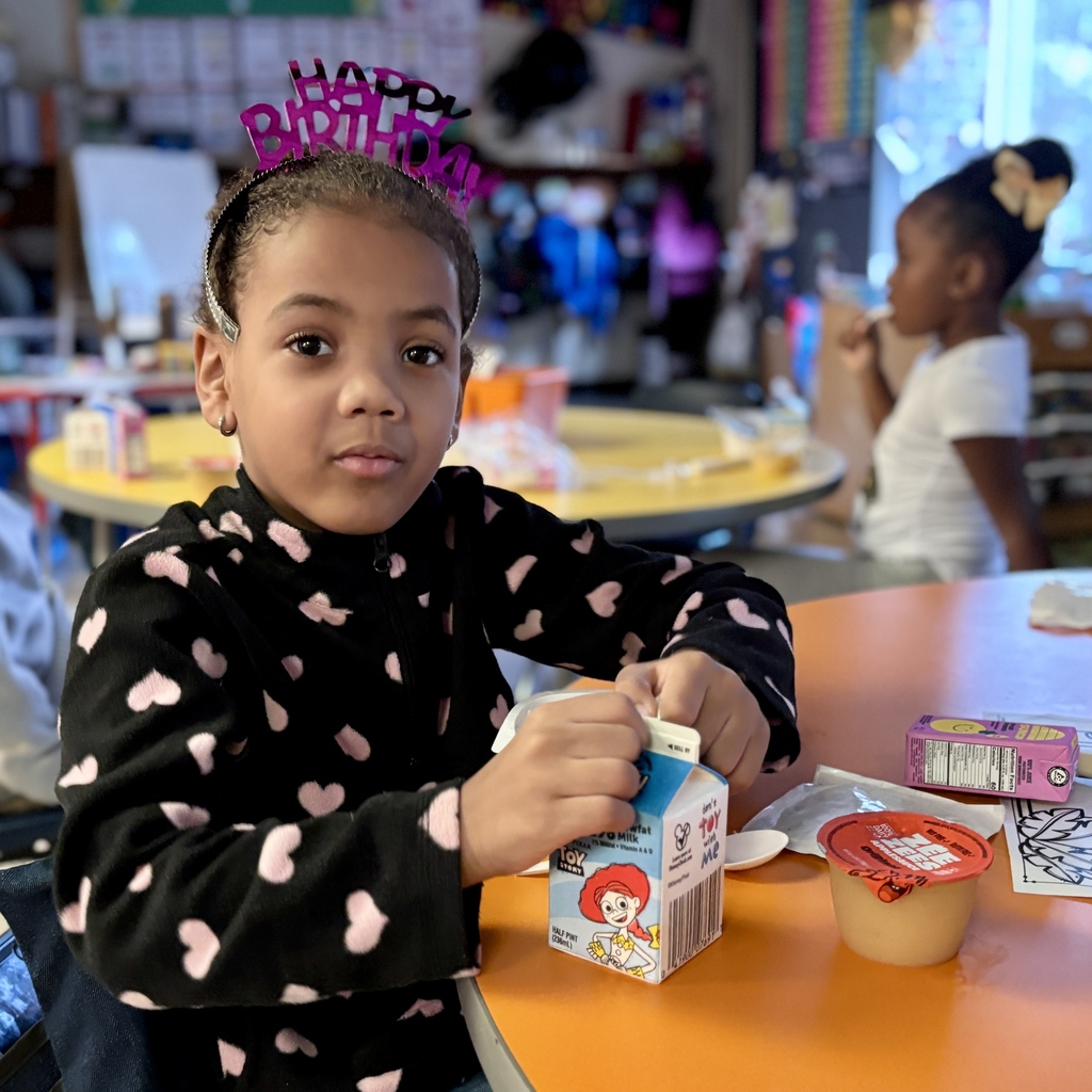 A young girl in a heart-printed jacket opens a Toy Story milk carton in a classroom.