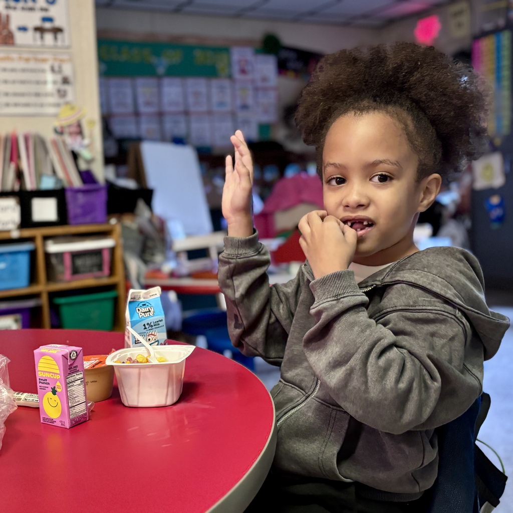 A child sits at a round red table in a classroom, eating a snack and raising one hand.