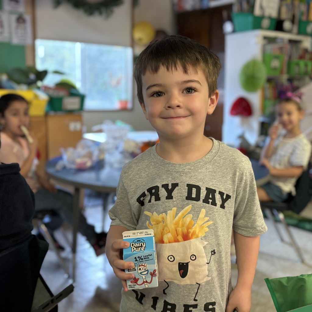A young boy with short brown hair smiles while holding a carton of milk in a classroom.