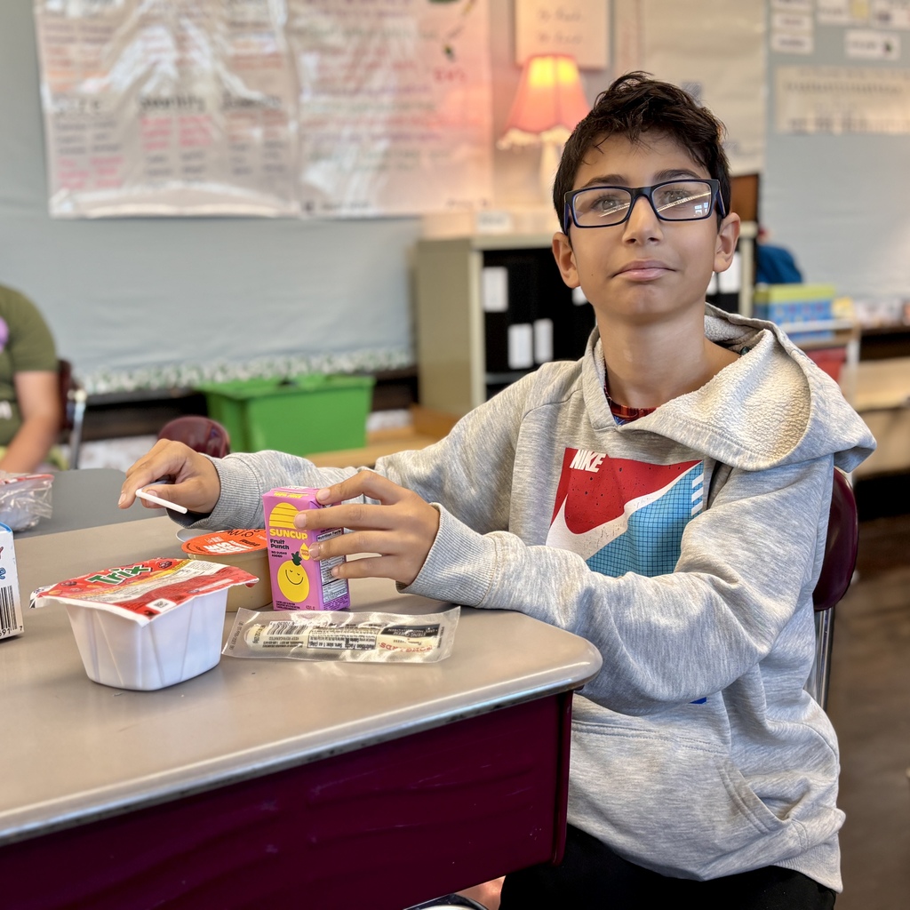 A young boy with glasses wearing a gray hoodie sits at a school desk, holding a juice box.