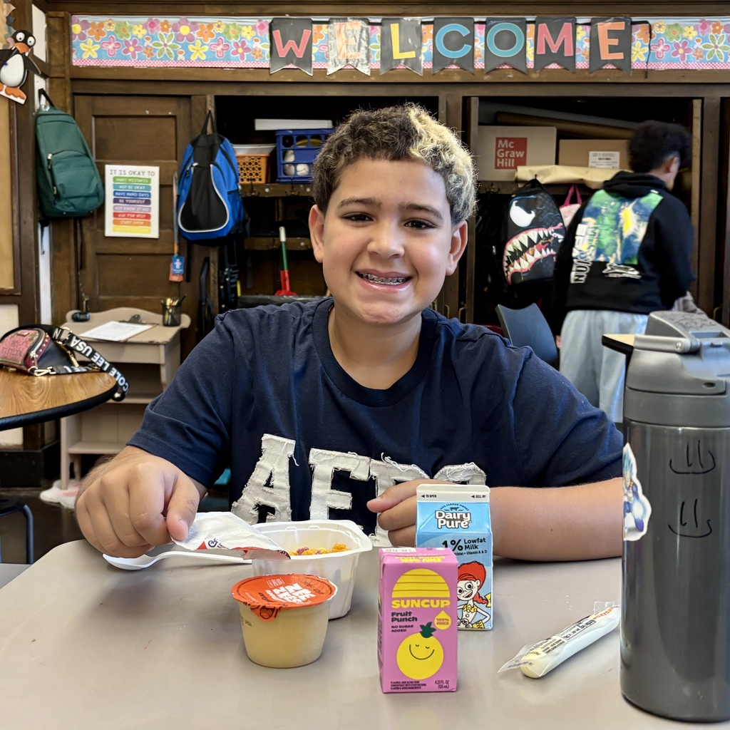 A smiling student sits at a desk in a classroom, eating lunch with milk, fruit punch, and cereal.