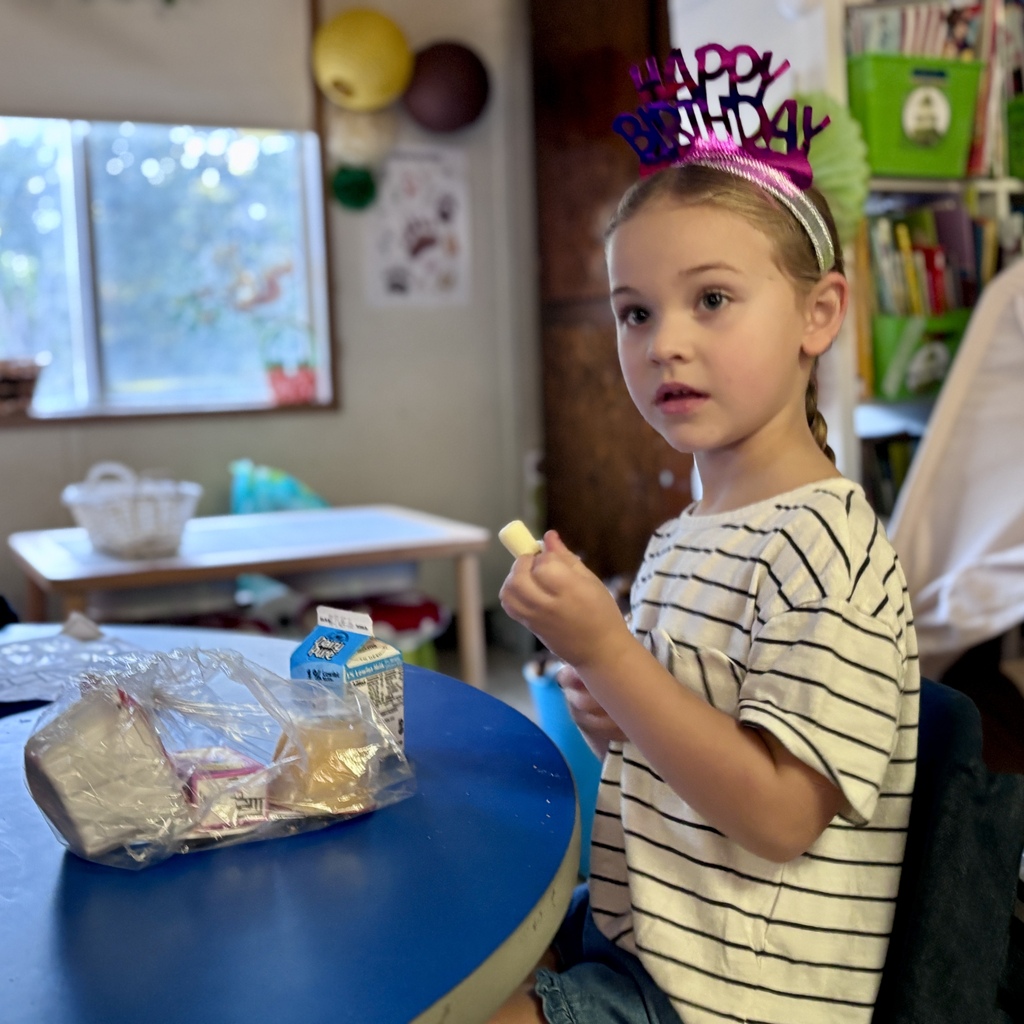 A child sits at a round red table in a classroom, eating a snack and raising one hand.