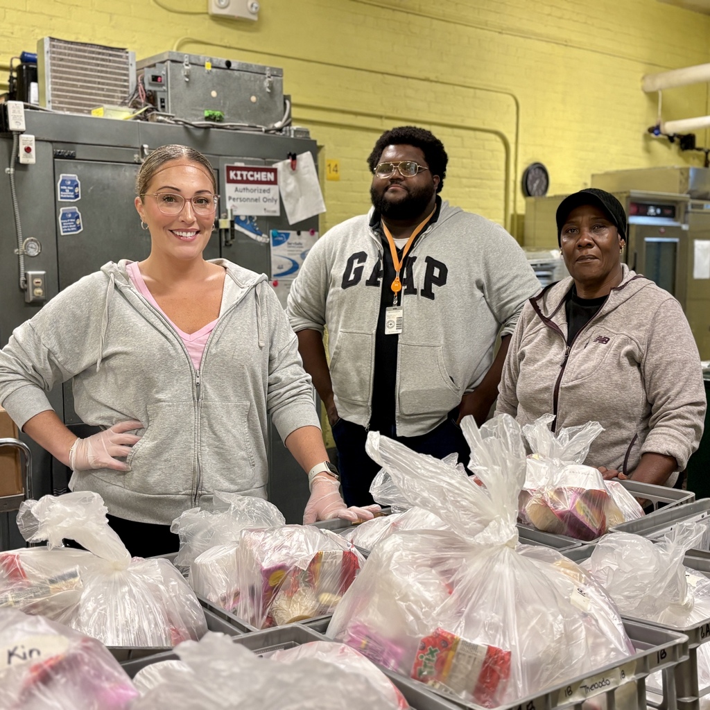 Three people pose behind crates of food bags in a commercial kitchen.